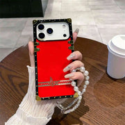 Phone case with red back and gold accents held by a hand with a pearl bracelet on a wooden table.