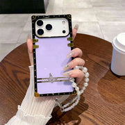 Square iPhone case with butterfly design and pearl strap held by a hand on a wooden table.