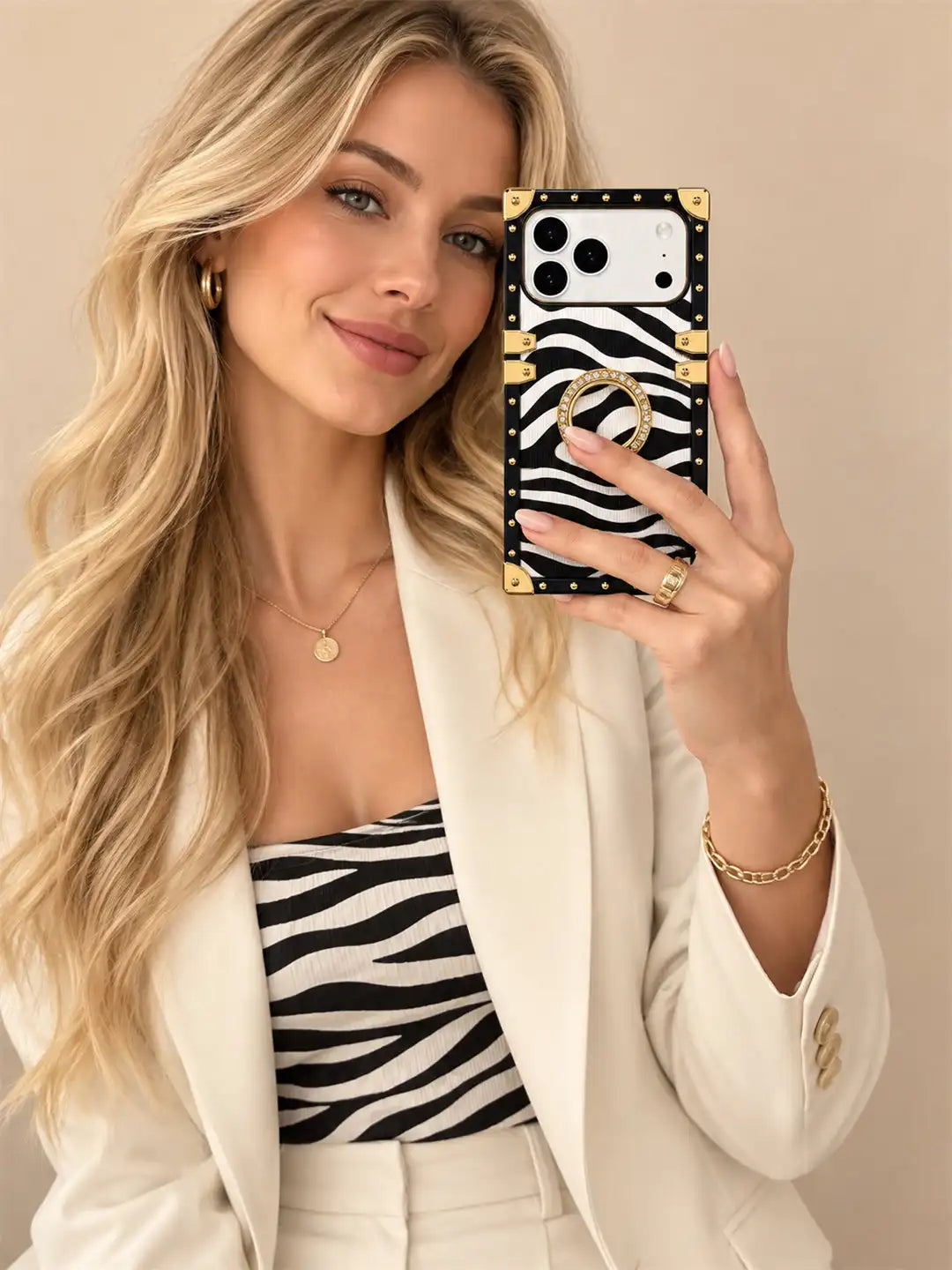 A woman takes a mirror selfie showing a phone case with a black and white zebra print, gold-tone studs, and a gold ring holder. She is wearing a cream blazer and a matching zebra-print top against a solid beige background.