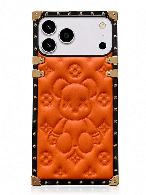An orange leather phone case featuring a teddy bear design and embossed monogram motifs, framed by a black border with gold-tone studs and corner protectors.
