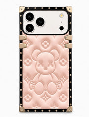 A luxury-style phone case featuring a pale pink embossed teddy bear design with classic monogram-style floral motifs, framed by a black border with gold-tone studs and corner protectors.