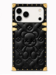 A luxury-style black phone case featuring a large embossed teddy bear motif in the center, surrounded by subtle monogram-style floral details. The case has a black leather-like finish with prominent gold-tone metal corner protectors and rivets, and a gold accent bar near the top.