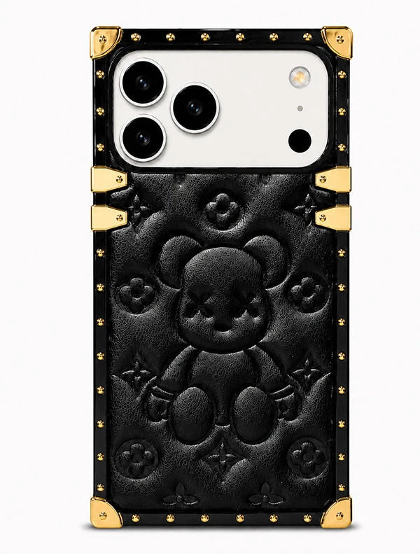 A luxury-style black phone case featuring a large embossed teddy bear motif in the center, surrounded by subtle monogram-style floral details. The case has a black leather-like finish with prominent gold-tone metal corner protectors and rivets, and a gold accent bar near the top.