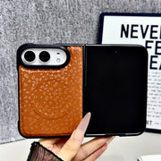 A hand holds a Samsung Galaxy Z Flip smartphone case that is open, showing a split design: a textured brown leather back panel on the left and a black panel on the right. The background features a gray textured wall and a decorative print.