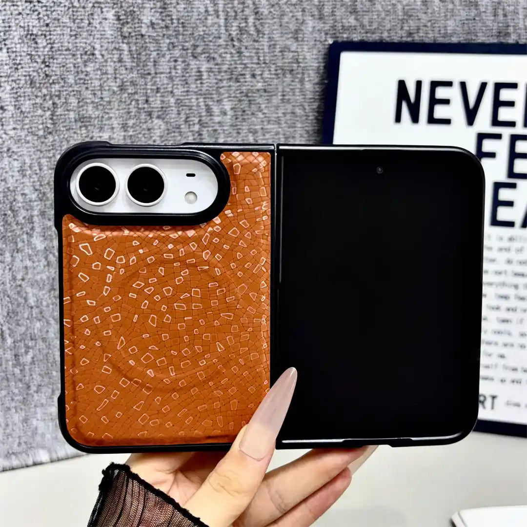 A hand holds a Samsung Galaxy Z Flip smartphone case that is open, showing a split design: a textured brown leather back panel on the left and a black panel on the right. The background features a gray textured wall and a decorative print.
