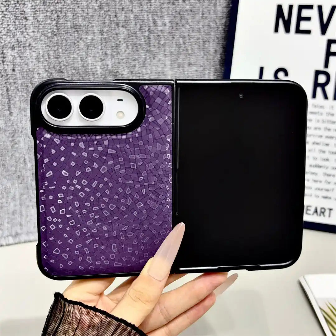 A hand holds a purple phone case with a mosaic tile pattern on the back of a black foldable smartphone. The background features a gray textured wall and a framed print with text.