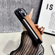 A hand holding a smartphone case with a textured tan-brown back and black bumper edges, photographed indoors against a gray fabric backdrop and a framed print.