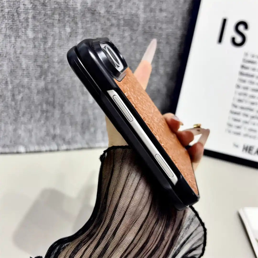 A hand holding a smartphone case with a textured tan-brown back and black bumper edges, photographed indoors against a gray fabric backdrop and a framed print.