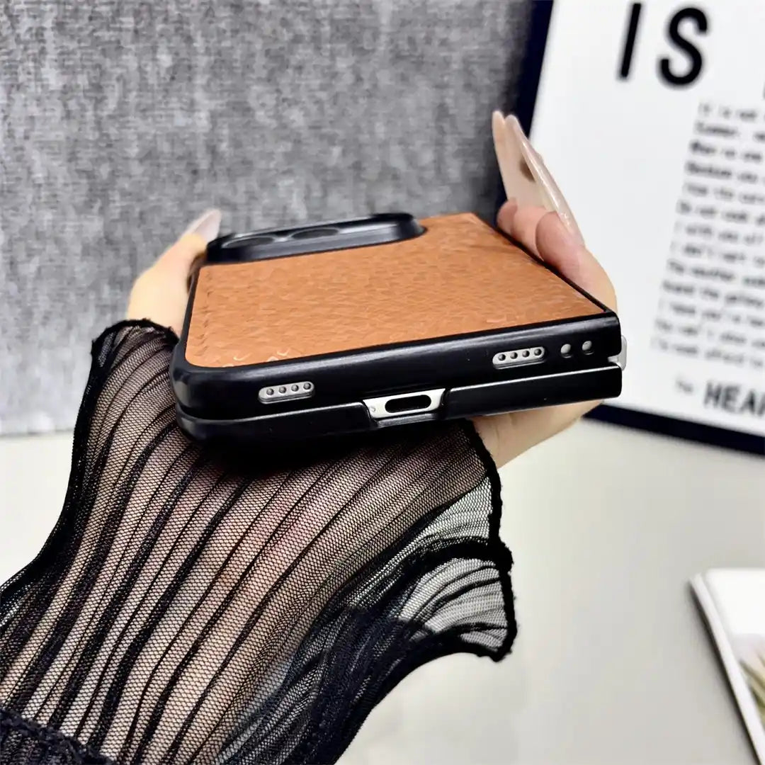 A hand holding a black smartphone case with a textured brown leather or faux-leather back panel, shown against a gray wall and framed print background.