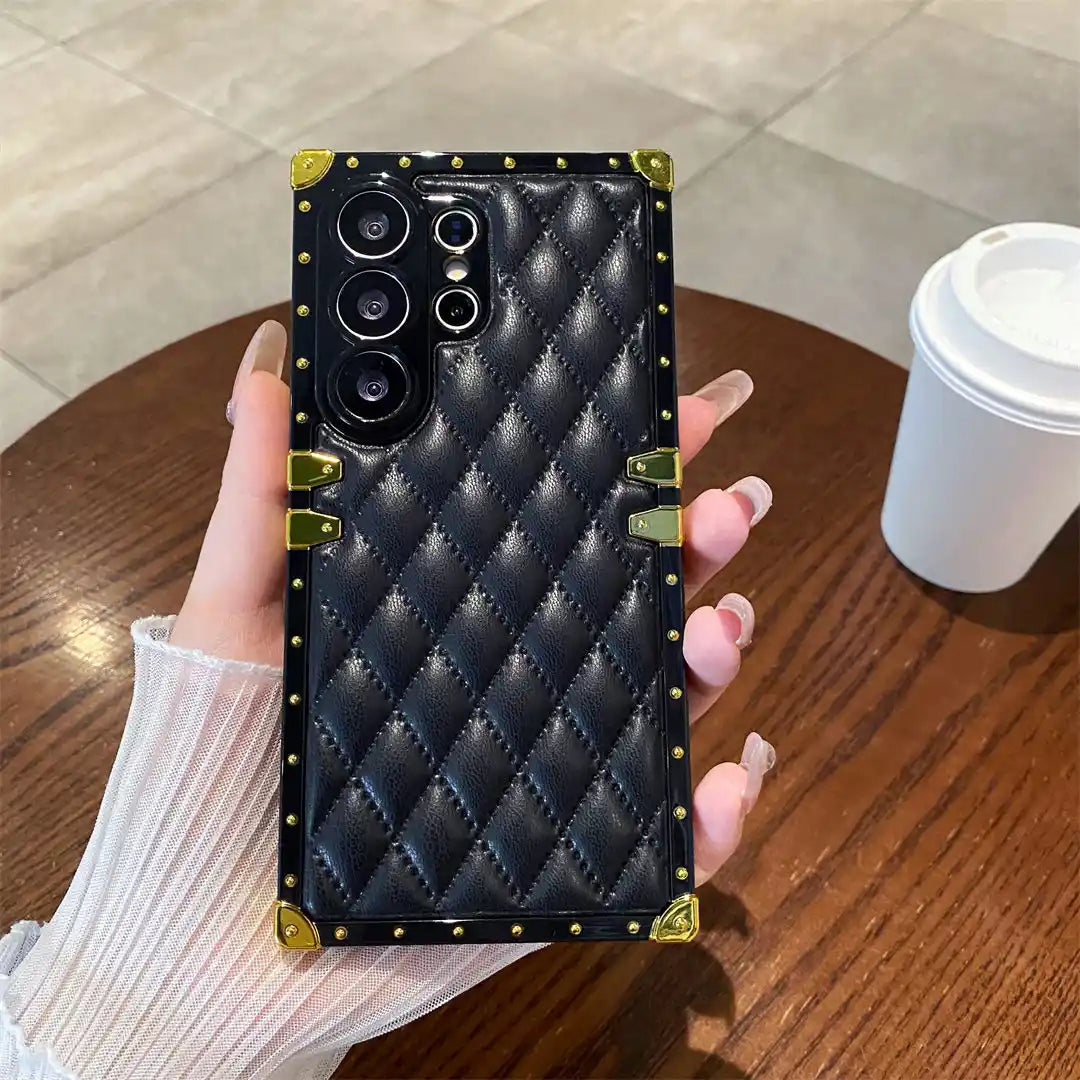 A hand holds a black quilted phone case with gold-tone metal corner protectors and studs, shown over a wooden table with a coffee cup in the background.