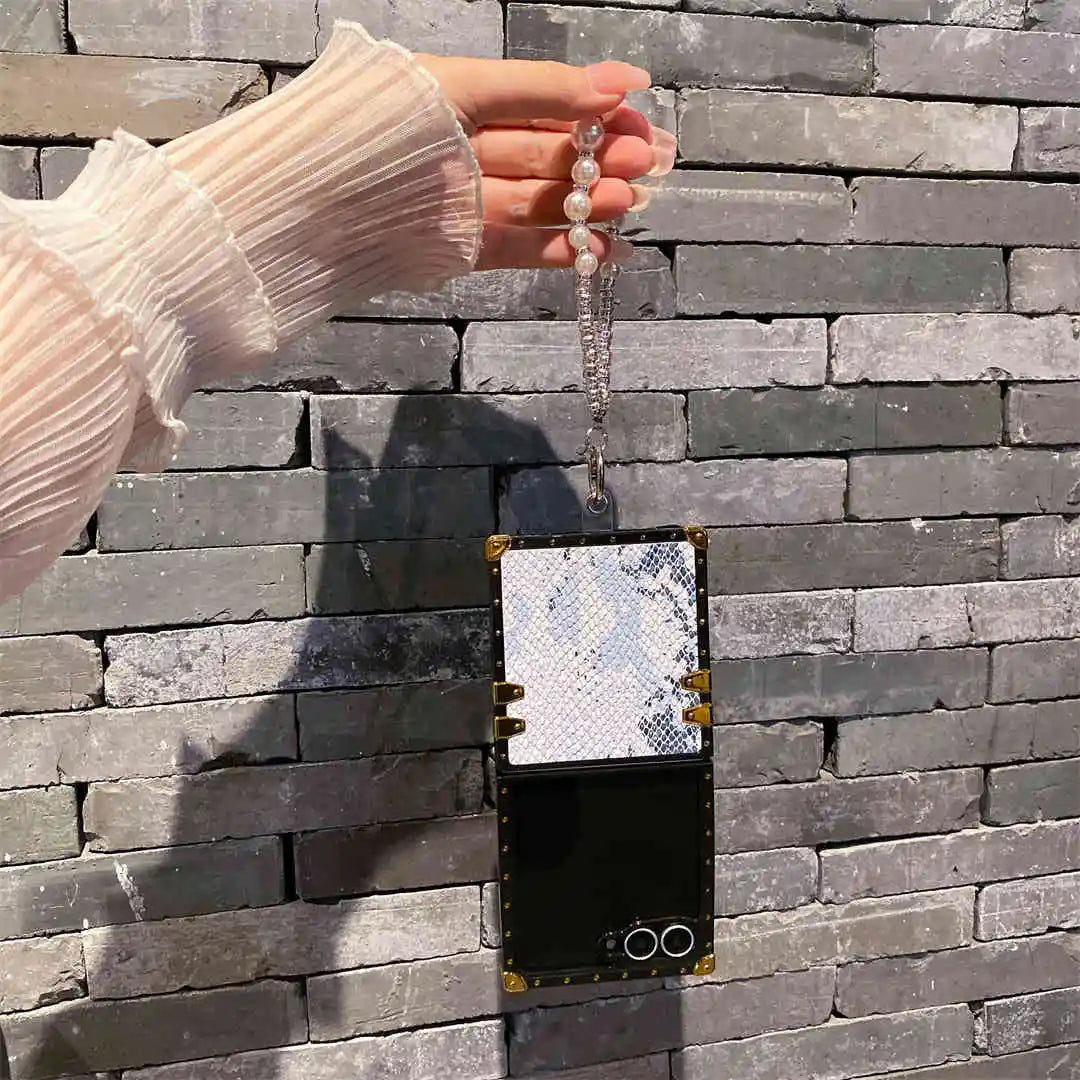 Hand holding a Galaxy Z flip 7 phone case with a decorative chain against a stone wall.