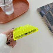 A hand holds a translucent neon yellow phone case, showing the bottom port openings, against a light tabletop with a drink tray and magazine in the background.