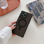 A hand wearing a white long-sleeve shirt holds a dark brown magnetic phone case with a circular MagSafe design, set against a light tabletop with a book and a glass of water in the background.