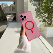 A hand wearing a white long-sleeve shirt holds a translucent pink phone case with a red MagSafe ring and red stylized accent on the back, photographed in a bright indoor setting with a window and greenery in the background.