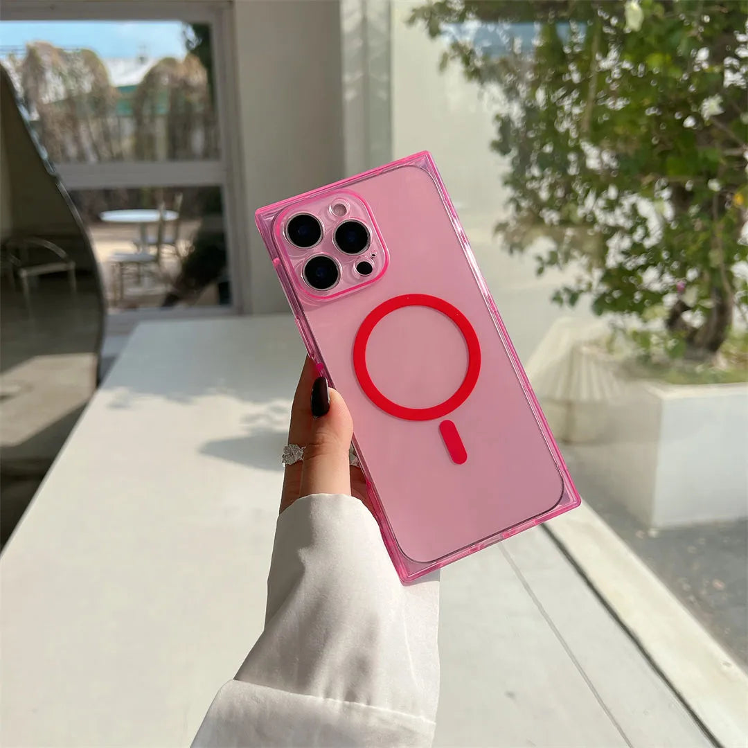 A hand wearing a white long-sleeve shirt holds a translucent pink phone case with a red MagSafe ring and red stylized accent on the back, photographed in a bright indoor setting with a window and greenery in the background.
