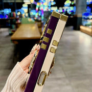 A close-up of a hand holding a purple and cream phone case with gold-tone metal accents and rhinestone embellishments, set against a blurred cafe background.