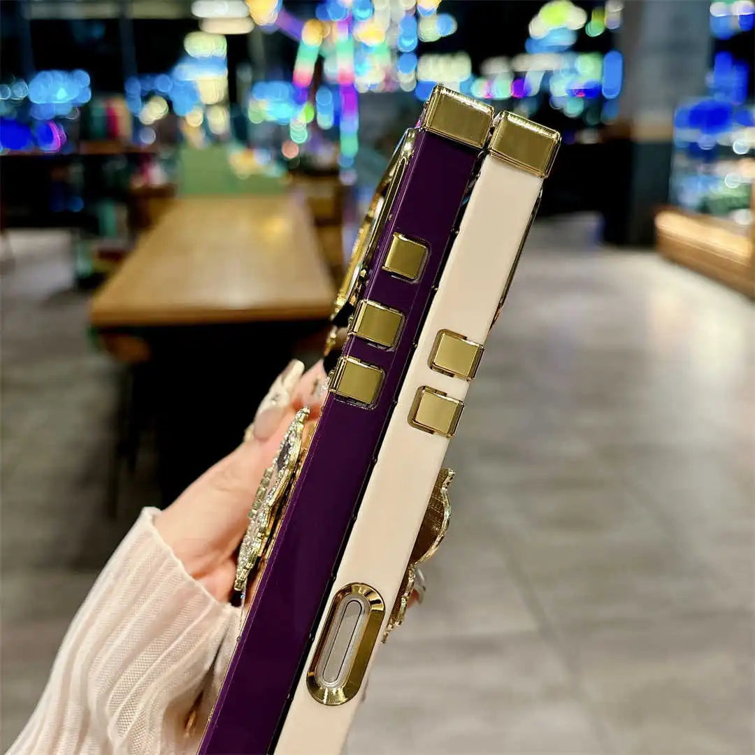 A close-up of a hand holding a purple and cream phone case with gold-tone metal accents and rhinestone embellishments, set against a blurred cafe background.