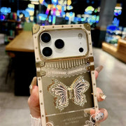 A hand holds a luxury gold-toned phone case for a foldable smartphone, featuring a large rhinestone-embellished butterfly design and decorative rivets, set against a blurred indoor background.
