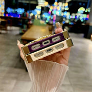 A hand holds two slim, rectangular electronic devices with gold-toned edges and decorative crystal-like detailing, shown against a blurred indoor background with colorful lights.