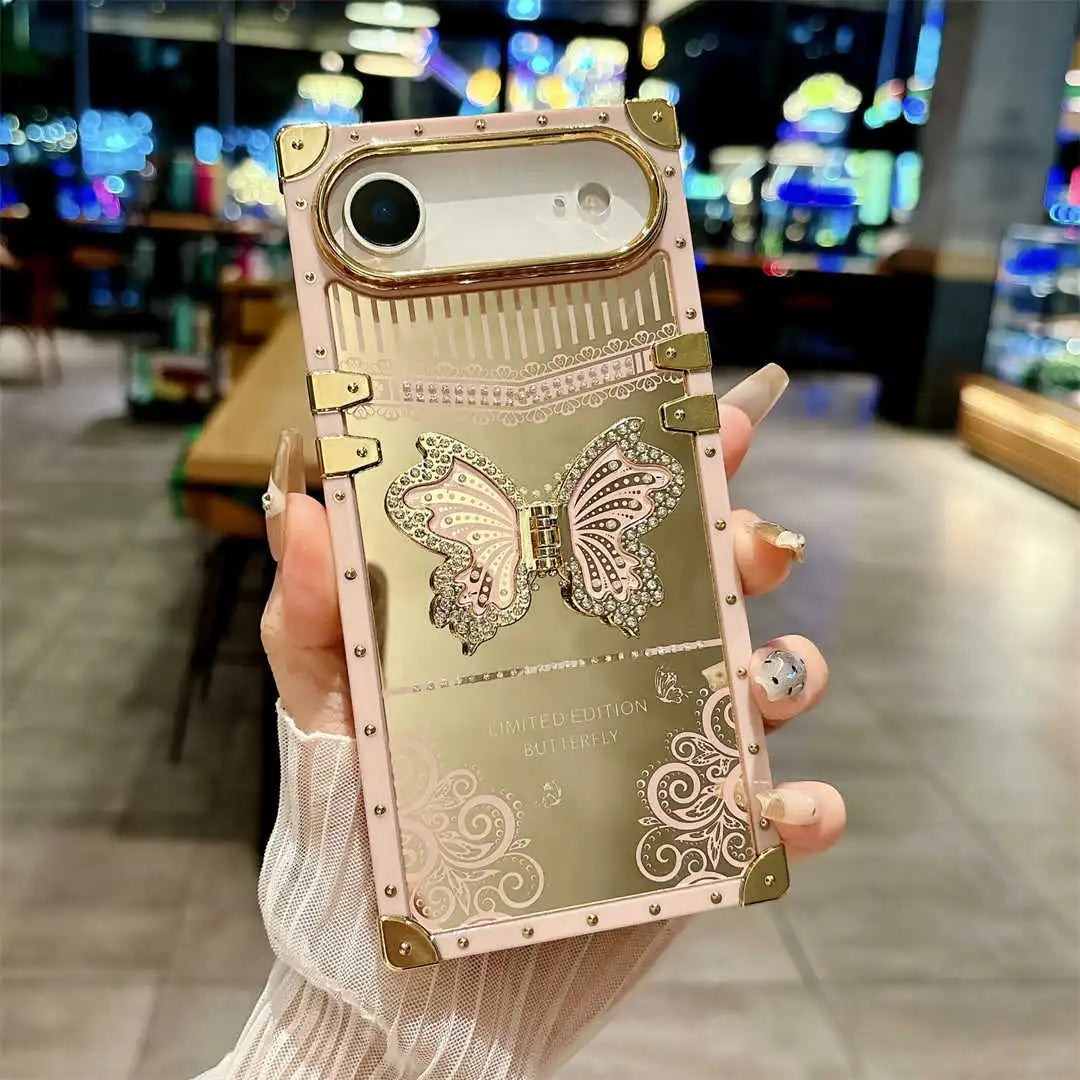 A hand holds a decorative phone case in a blurred indoor setting. The case is metallic rose gold with gold-toned corner protectors, rivet details, and a large glittery butterfly design in the center.