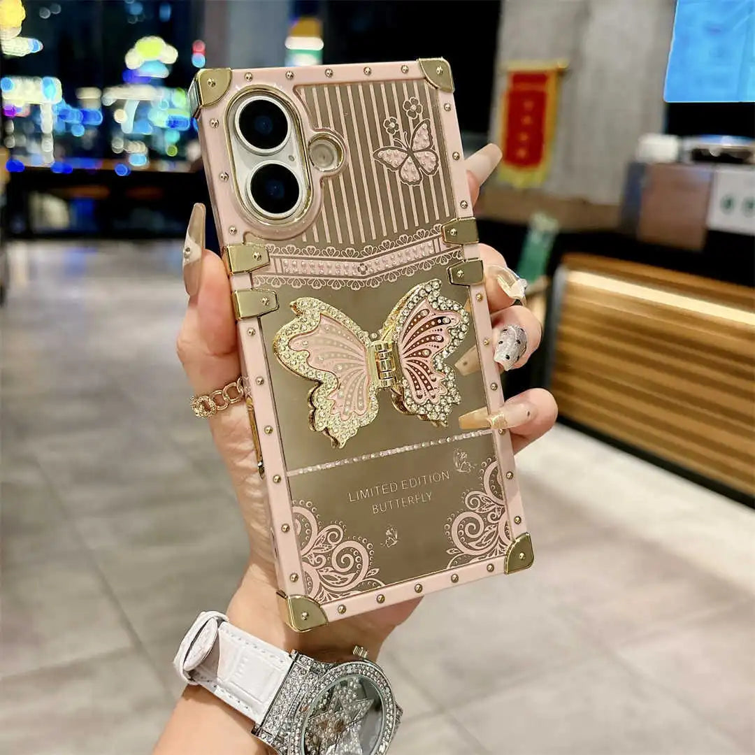 A hand holds a decorative smartphone case in a blurred indoor setting. The case is metallic rose gold with embossed butterfly designs, gold-tone corner protectors, and ornate scrollwork details.