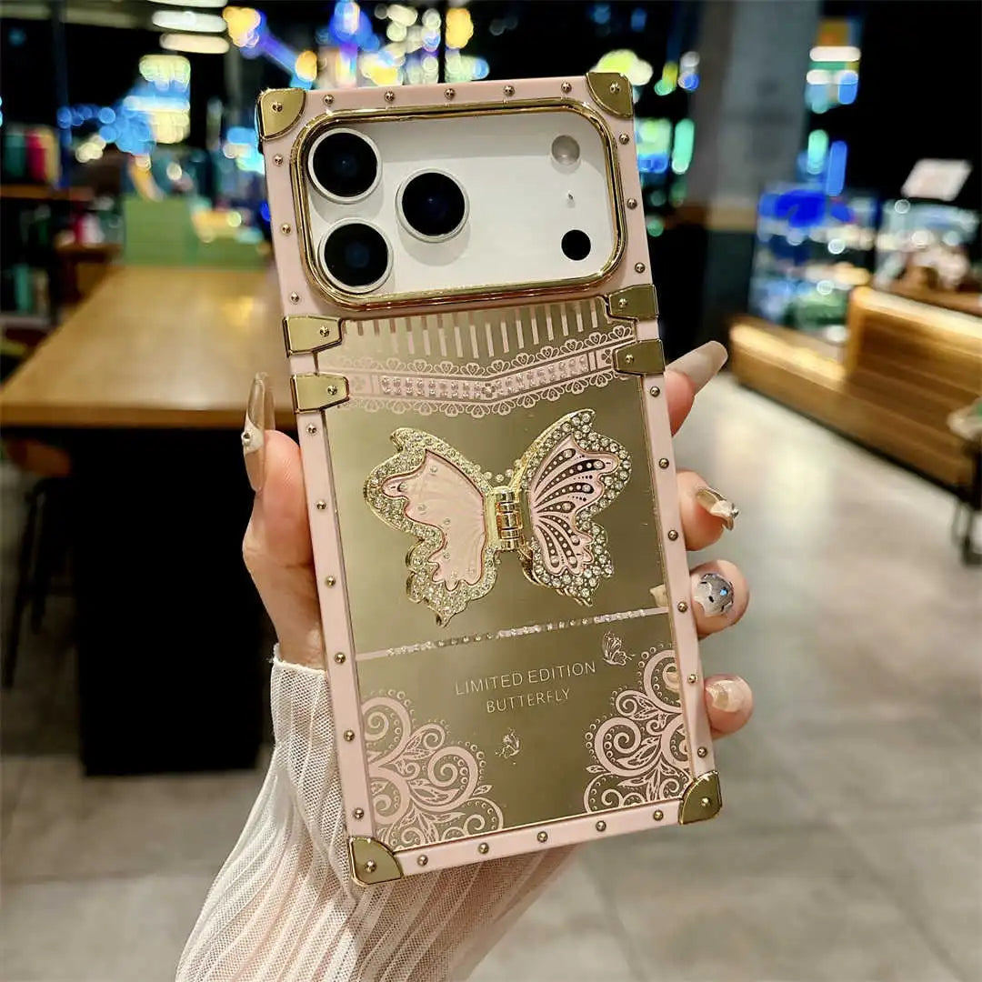 A hand holds a metallic rose gold and gold phone case featuring a large butterfly design, decorative scrollwork, and gold-tone rivets, with a blurred cafe interior in the background.