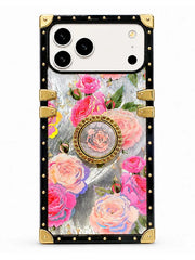 A protective phone case with a luxury design, featuring a black border with gold-tone rivets and corner guards, a central floral print of pink and peach roses, and a circular rose medallion on a light gray textured background.