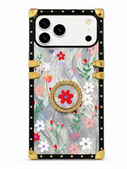 A protective phone case with a vintage-style trunk design, featuring a black studded border, gold corner protectors, and a clear back decorated with a colorful floral pattern and a central gold-and-red medallion ring.