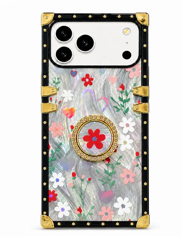 A protective phone case with a vintage-style trunk design, featuring a black studded border, gold corner protectors, and a clear back decorated with a colorful floral pattern and a central gold-and-red medallion ring.