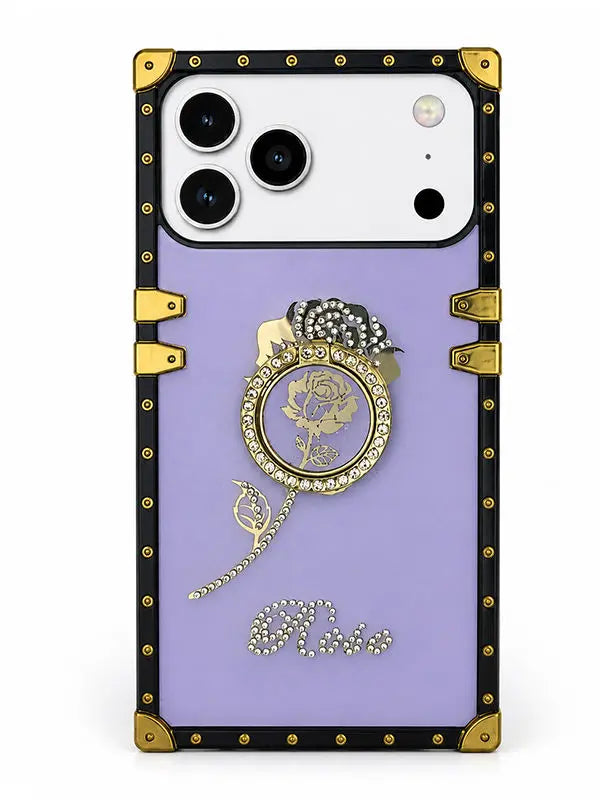 A luxury-style phone case with a lavender purple backplate, featuring a black and gold faux-leather trim with gold-tone studs and corner protectors. The center is decorated with a gold rose medallion, a rhinestone-embellished black bow, a dangling gold chain, and the word 'Elvii' in rhinestone script at the bottom.