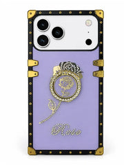 A luxury-style phone case with a lavender purple backplate, featuring a black and gold faux-leather trim with gold-tone studs and corner protectors. The center is decorated with a gold rose medallion, a rhinestone-embellished black bow, a dangling gold chain, and the word 'Elvii' in rhinestone script at the bottom.