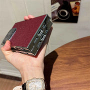 Hand holding a small rectangular device with a maroon surface and metallic accents, on a wooden surface.