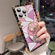 A hand holding a smartphone case with a pink marble and glitter geometric design, black border, gold corner accents, and a built-in ring stand, photographed in a real indoor setting with blurred chairs and lights in the background.