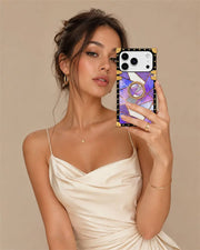 A woman in a cream satin dress takes a mirror selfie showing off a phone case. The case features a black border with gold studs, a gold ring handle, and a purple and white marble pattern.