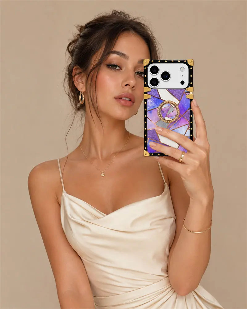 A woman in a cream satin dress takes a mirror selfie showing off a phone case. The case features a black border with gold studs, a gold ring handle, and a purple and white marble pattern.