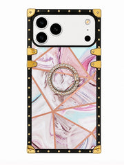 A protective phone case with a black border, gold corner protectors and rivets, a geometric rose gold line pattern, and a pink and white marble swirled center. It features a built-in ring holder in the middle adorned with clear rhinestones.