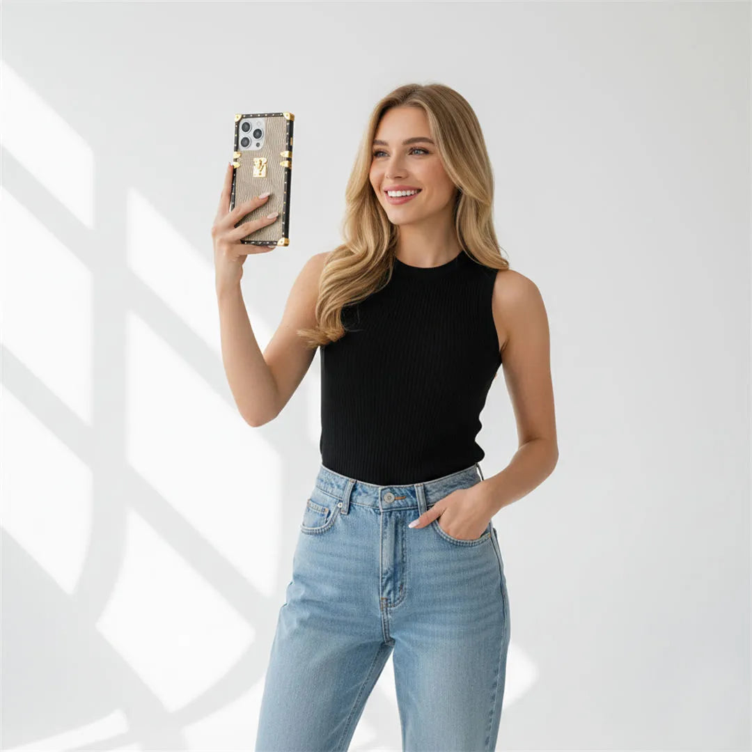 Woman taking a selfie with a phone, wearing a black sleeveless top and light blue jeans, on a white background.