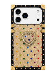 A protective phone case with a luxury trunk design, featuring a gold glitter body decorated with small multicolored hearts, a central rhinestone-embellished heart stand, and a black border with gold-tone studs and corner protectors.