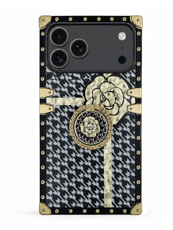 A luxury-style phone case featuring a black and white houndstooth pattern, gold-tone rivets, a large cream-colored camellia flower, and a central circular rose medallion, shown on a plain white background.