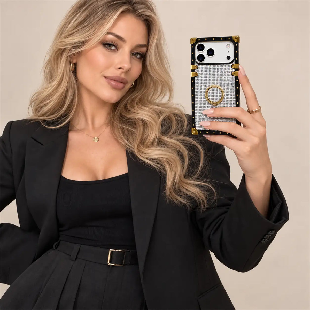 A woman in a black blazer and top takes a mirror selfie showing a phone case. The case is black with gold corner accents, a silver rhinestone-look center, and a gold ring stand.