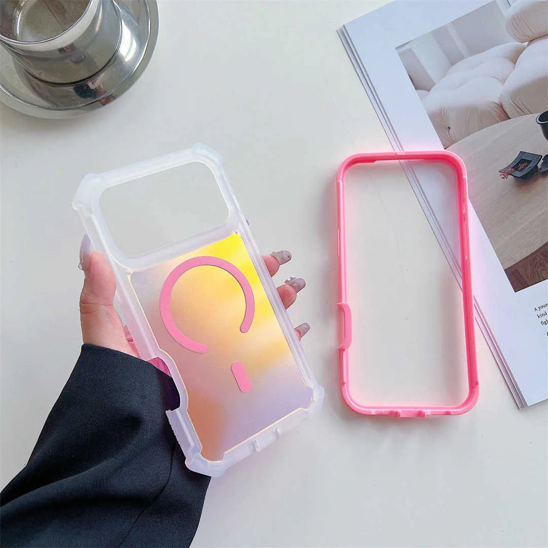 A hand holds a clear phone case with a pink and yellow gradient insert, while a separate pink bumper frame lies beside it on a light tabletop. An open magazine and a metallic cup are partially visible in the background.