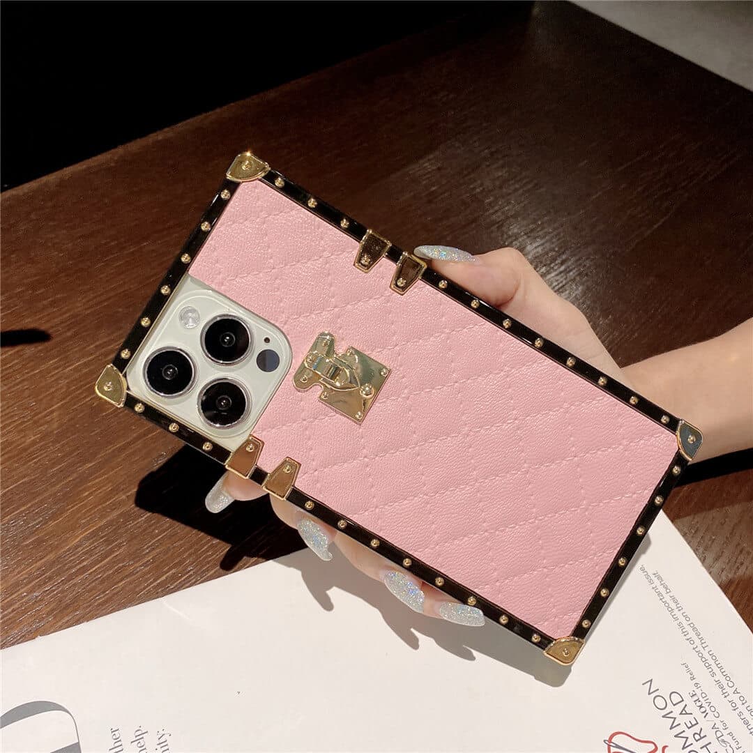 A pink quilted phone case with gold-tone metal studs and corner protectors, held by a hand over a dark wooden table.