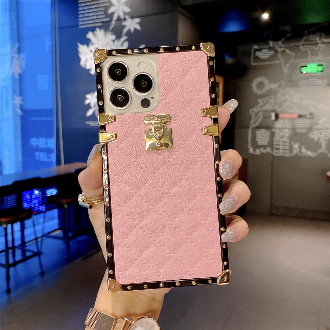 A hand holds a pink textured phone case with a quilted diamond pattern, gold-tone hardware, and a decorative clasp, shown against a blurred indoor background.