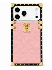 A pink textured phone case designed to look like a luxury trunk, featuring a black border with gold-tone studs, corner protectors, and a central gold clasp lock, shown on a white smartphone.