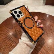 A hand wearing a white long-sleeve top holds a luxury-style phone case with a quilted tan leather back, black trim, gold-tone rivets, and a circular magnetic ring, set against a wooden table background.