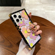A hand holds a smartphone case decorated with a pastel floral pattern, featuring gold-tone metal corner protectors and a built-in ring stand. The background shows a wooden table and tiled floor.