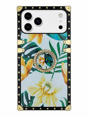 A protective phone case with a tropical floral print featuring yellow hibiscus and green monstera leaves on a white background. The case has a black border with gold-tone rivets and corner protectors, and a circular crystal-embellished ring stand in the center.