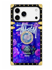 A protective phone case featuring a vibrant purple and blue floral design with a prominent white Stussy logo, a circular rose medallion, and 'London Paris' text. The case has a black border with gold rivets and corner protectors, shown on a white background.