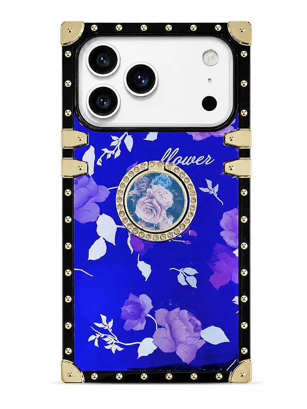 A luxury-style phone case with a royal blue floral print, black border, gold-tone metal corner protectors and rivets, and a central circular mirror accent.