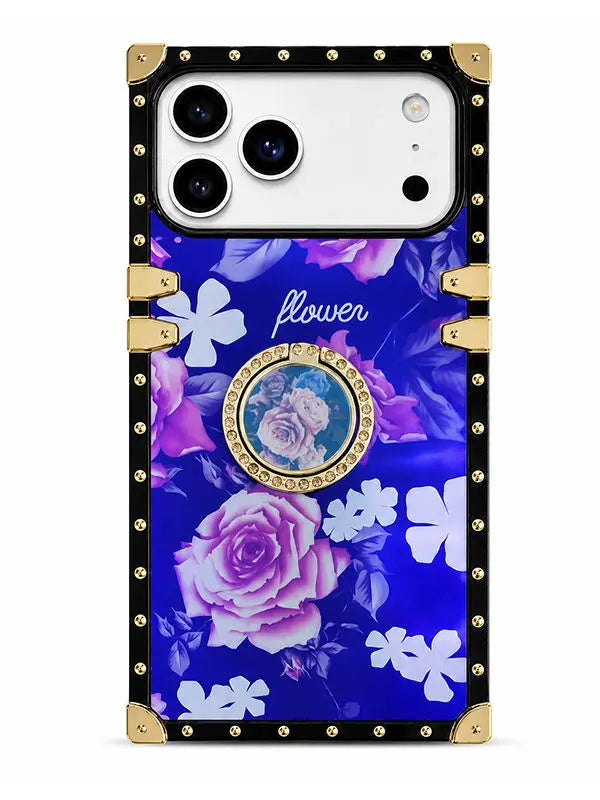 A protective phone case with a royal blue floral print, featuring pink and purple roses, white flowers, a central circular photo frame, and a black border with gold-tone rivets and corner protectors.